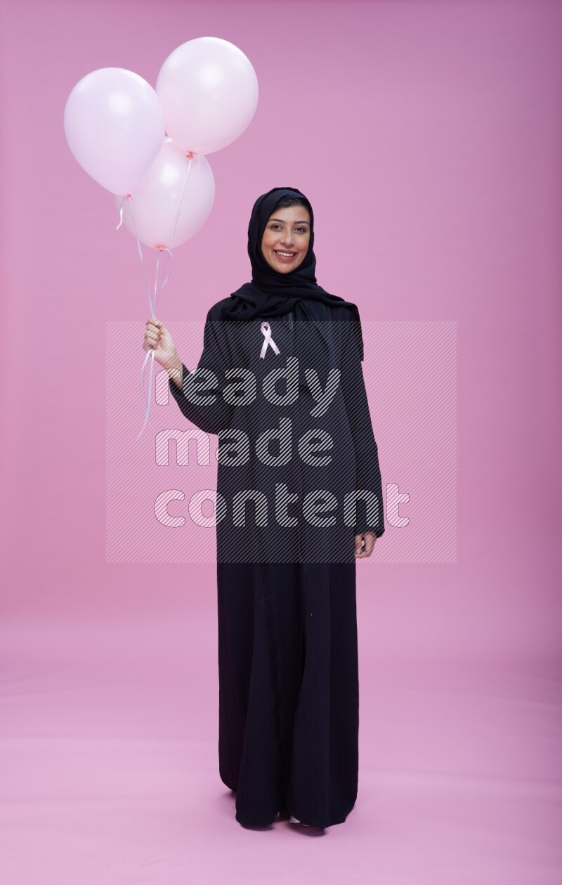 Saudi woman wearing pink ribbon on Abaya standing holding balloons on plain pink background