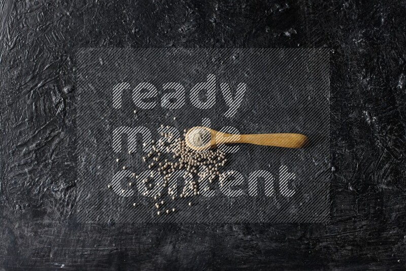 A wooden spoon full of white pepper powder with white pepper beads on textured black flooring
