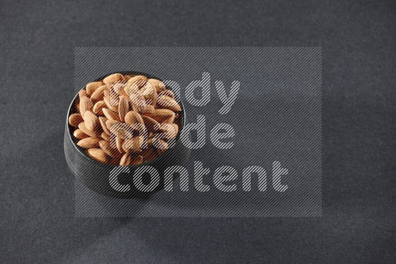 A black pottery bowl full of peeled almonds on a black background in different angles