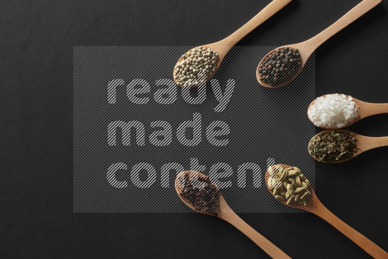wooden spoons filled with white peppers, cloves, cardamom, salt, black peppers and basil on black flooring