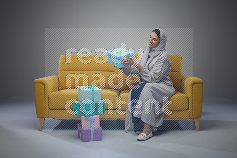 A Saudi woman wearing a light gray Abaya and head scarf sitting on a yellow sofa holding a gift box beside multi color gift boxes eye level on a grey background