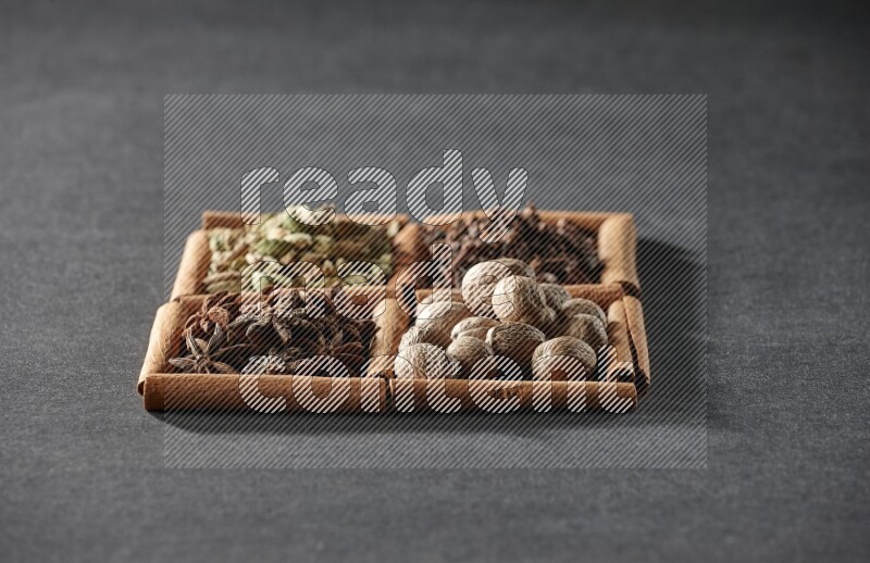 4 مربعات من عيدان القرفة ممتلئة بحب الهيل واليانسون النجمي وجوزة الطيب والقرنفل على أرضية سوداء