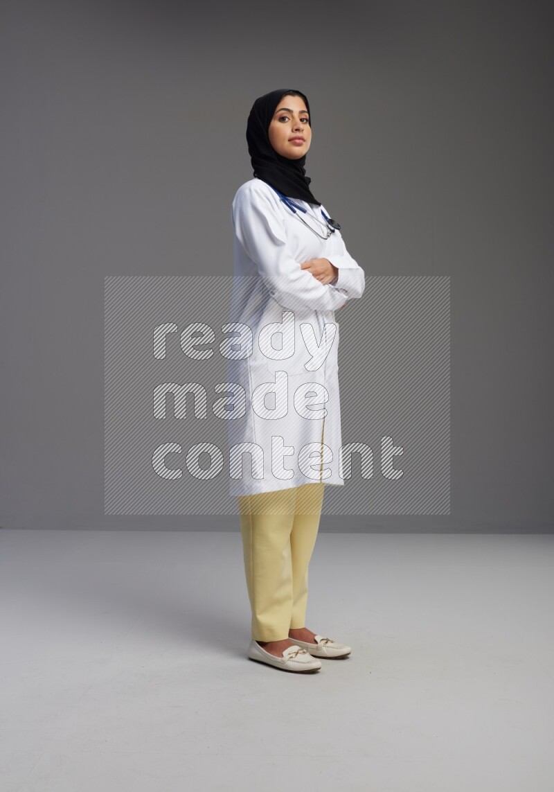 Saudi woman wearing lab coat with stethoscope standing with crossed arms on Gray background