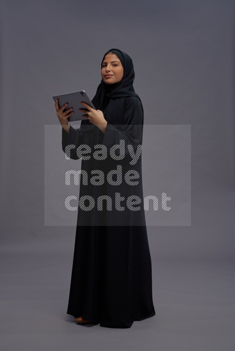 Saudi woman wearing Abaya standing working on tablet on gray background