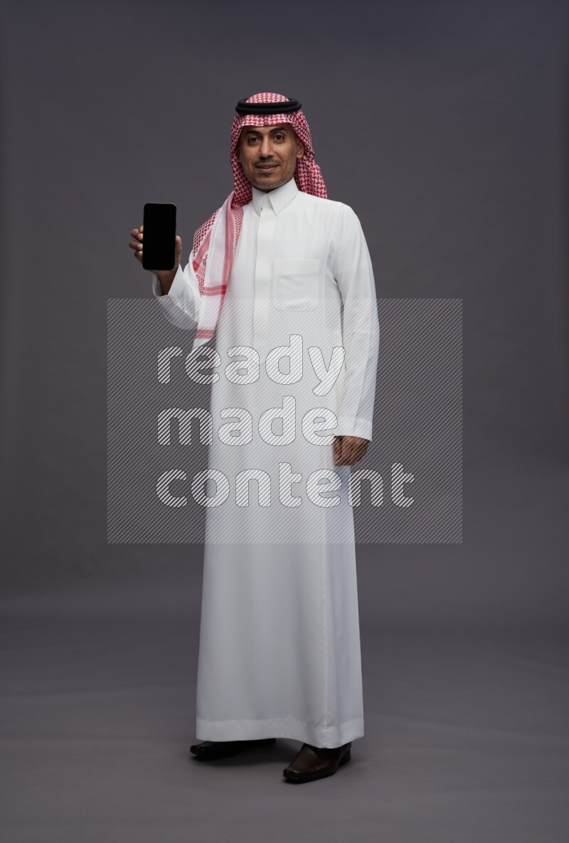Saudi man wearing thob and shomag standing showing phone to camera on gray background