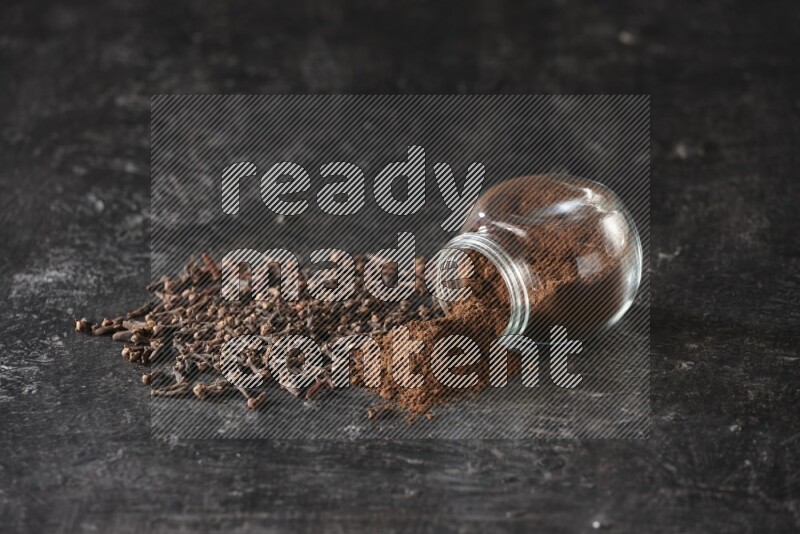 A flipped glass spice jar full of cloves powder and powder came out of it with cloves spread on textured black flooring