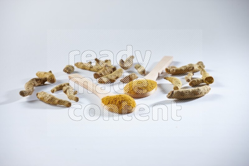 2 wooden spoons full of turmeric powder with dried turmeric fingers beside it on white flooring