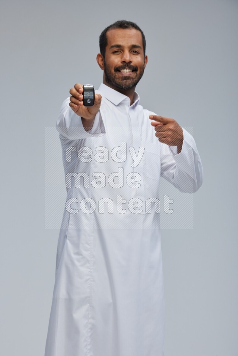 Saudi man wearing thob and shomag standing holding car key on gray background
