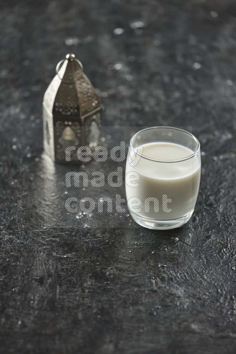 A silver lantern with different drinks, dates, nuts, prayer beads and quran on textured black background