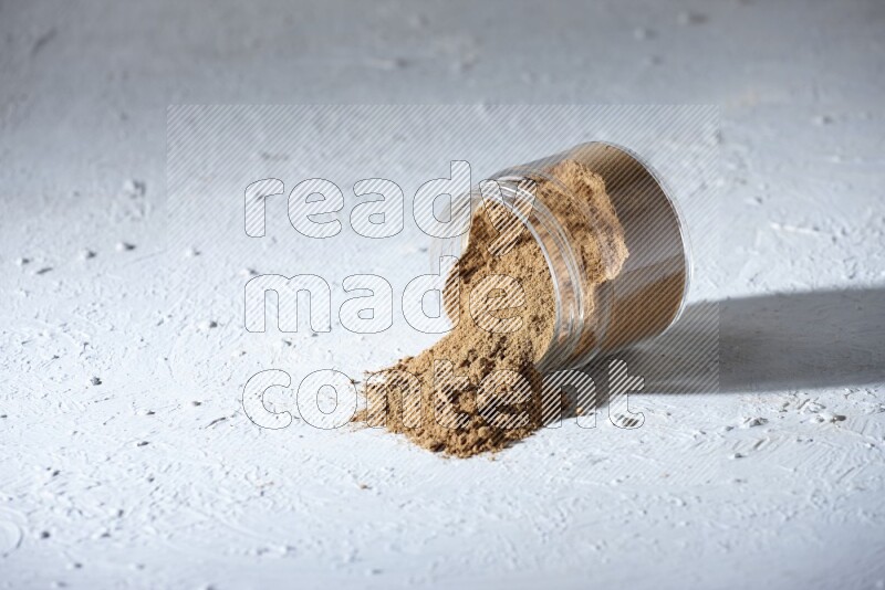 A flipped glass jar full of allspice powder and powder spilled out of it on a textured white flooring