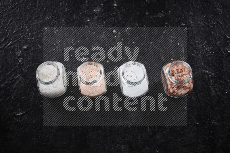 4 glass jars filled with table salt, coarse sea salt, fine himalayan salt and coarse himalayan salt on black background