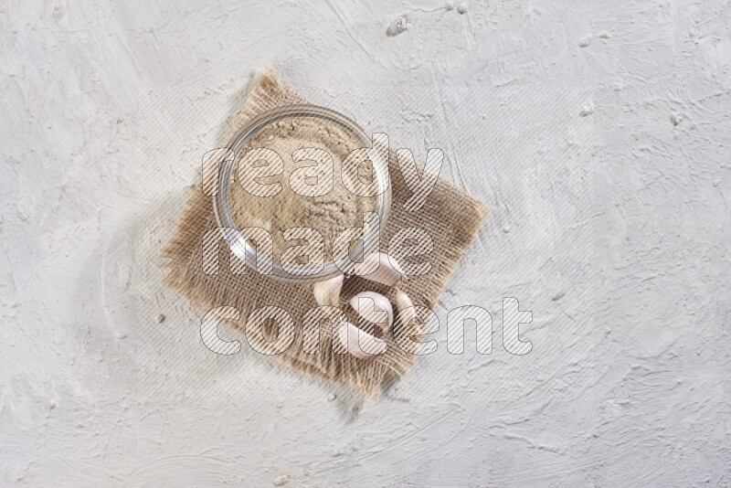 A glass bowl full of garlic powder placed on burlap fabric with garlic cloves on a textured white flooring