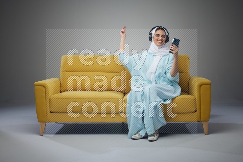 A Saudi woman wearing a light blue Abaya and a white head scarf sitting on a yellow sofa and holding an iPad while wearing headphone eye level on a grey background