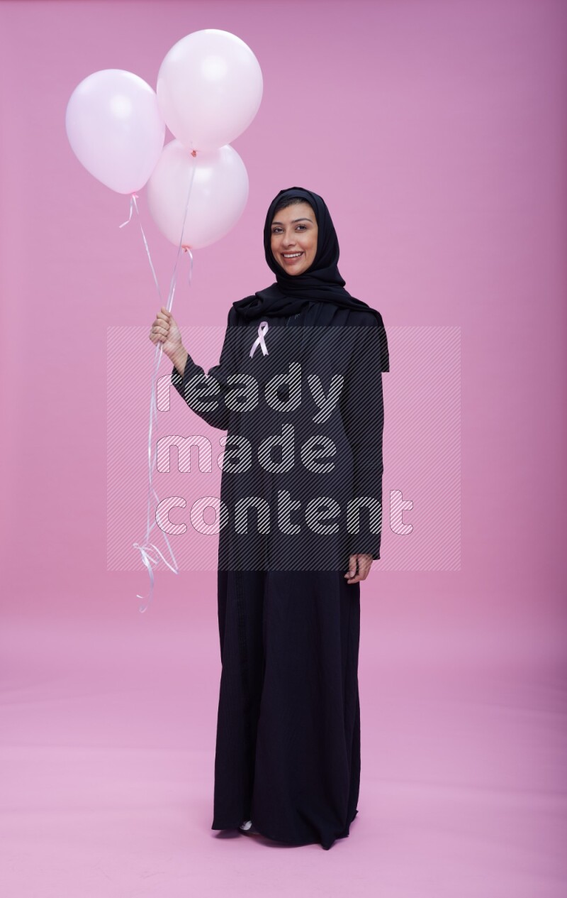 Saudi woman wearing pink ribbon on Abaya standing holding balloons on plain pink background