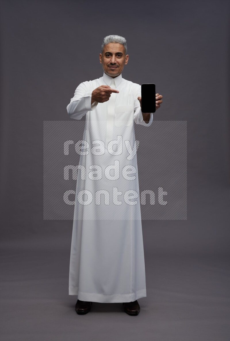Saudi man wearing thob standing showing phone to camera on gray background