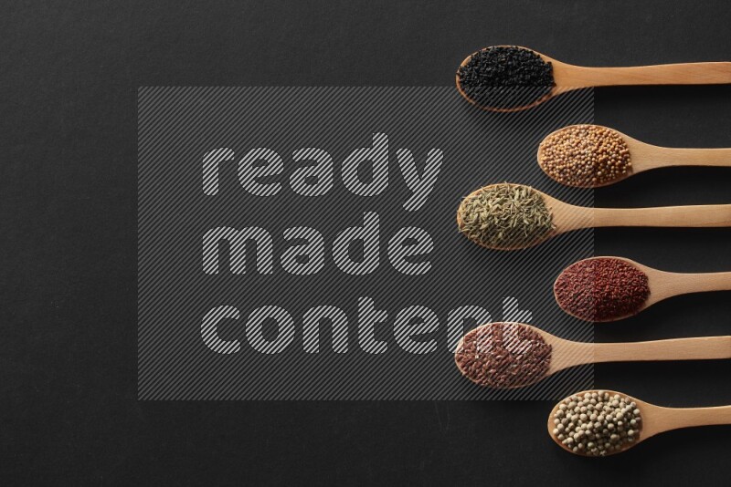 wooden spoons filled with white peppers, mustard seeds, black seeds, garden cress, cumin and flax on black flooring