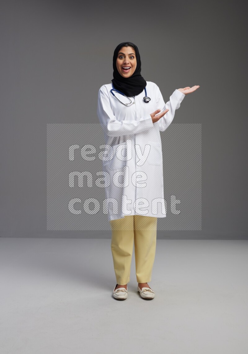 Saudi woman wearing lab coat with stethoscope standing interacting with the camera on Gray background