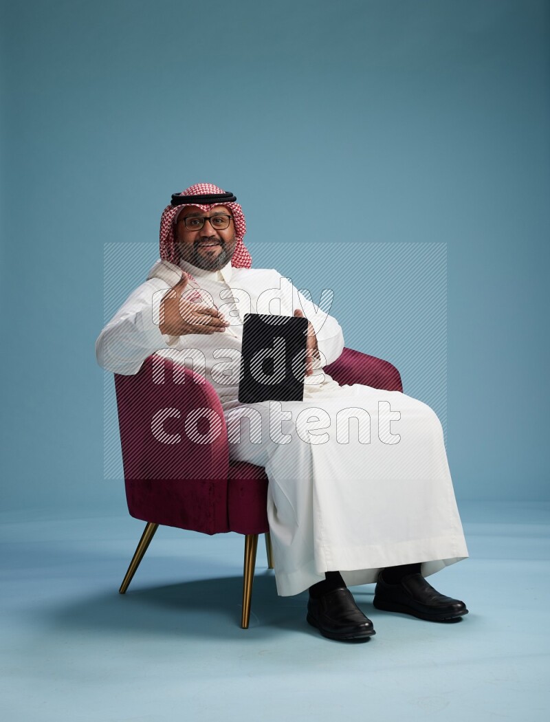 Saudi Man with shimag sitting on chair working on tablet on blue background