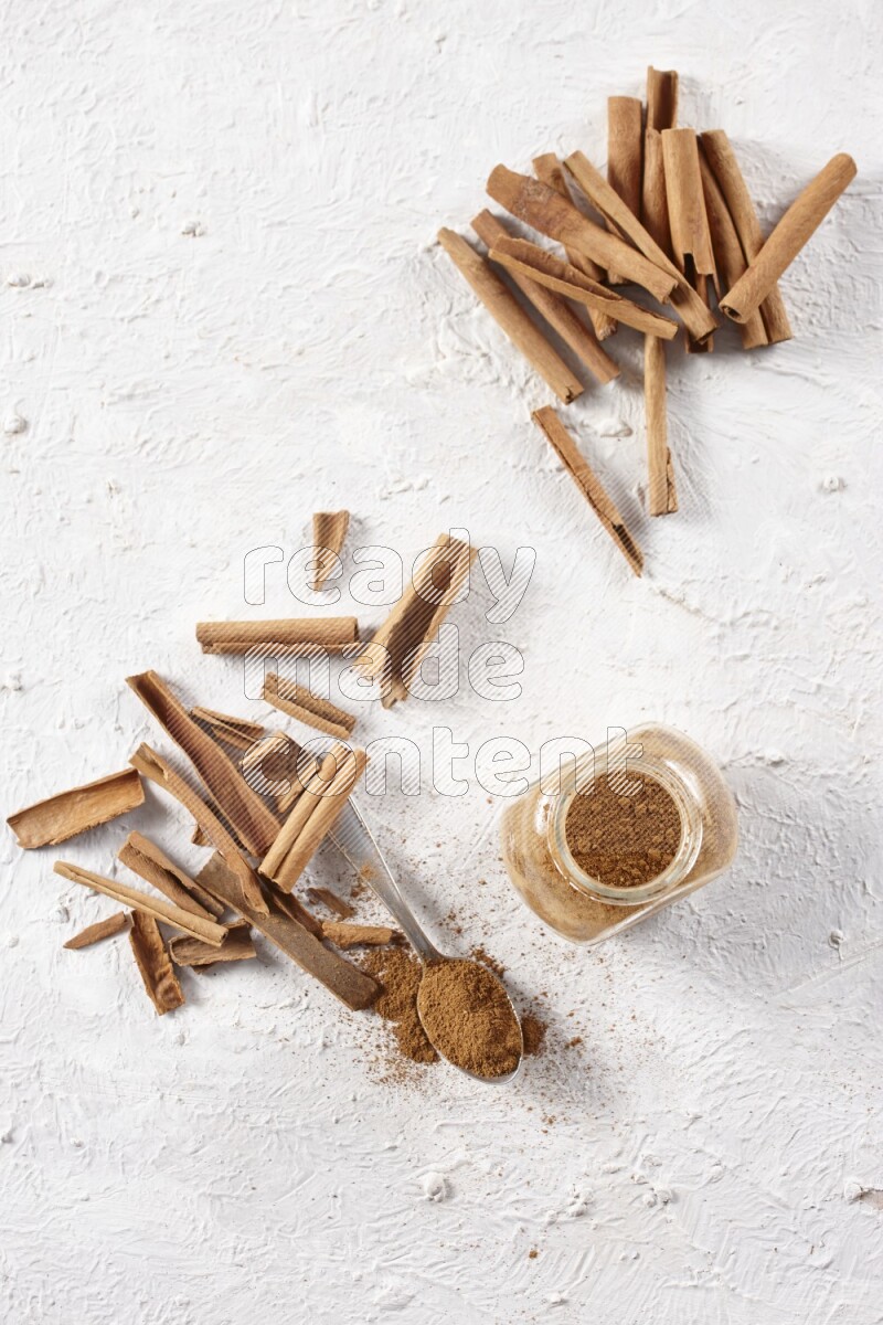 Herbal glass jar full cinnamon powder and a metal spoon surrounded by cinnamon sticks on a white background