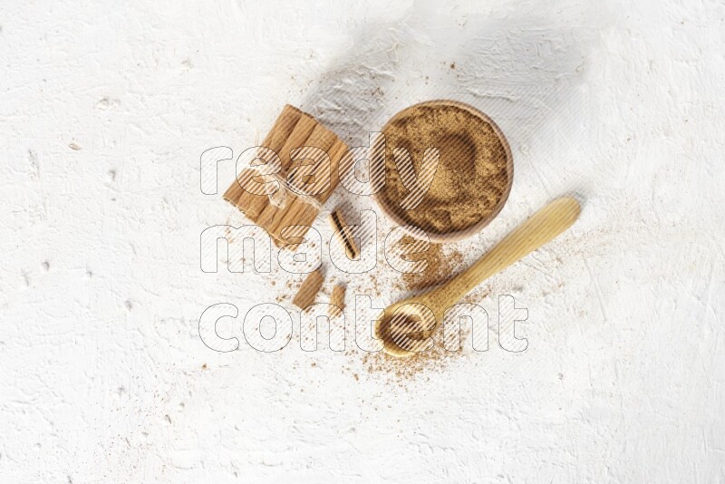 Cinnamon sticks stacked and bounded beside a wooden bowl full of cinnamon powder and a wooden spoon full of powder on white background