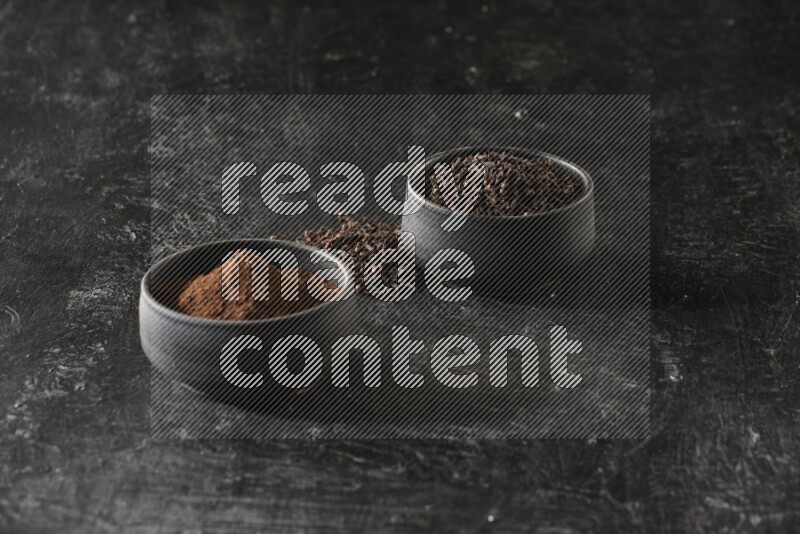2 Black pottery bowls full of cloves and the other full of cloves powder on textured black flooring