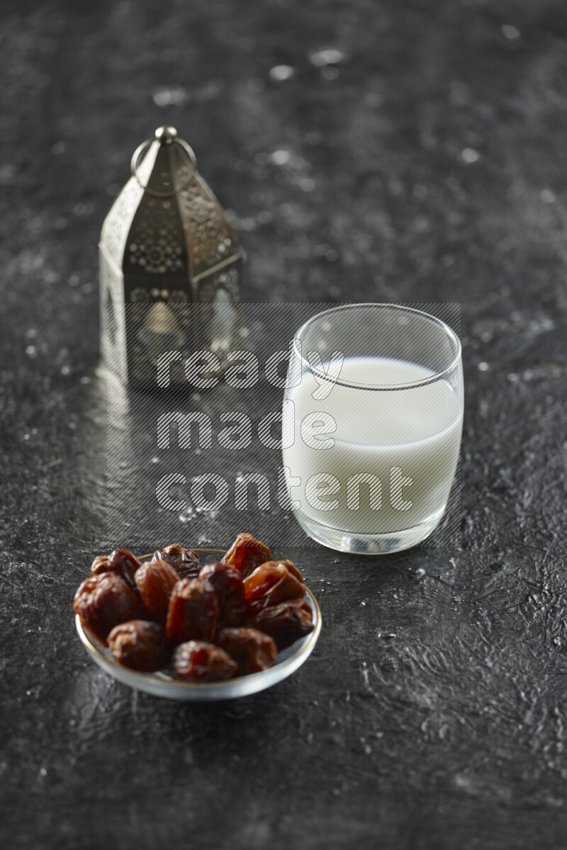 A silver lantern with different drinks, dates, nuts, prayer beads and quran on textured black background