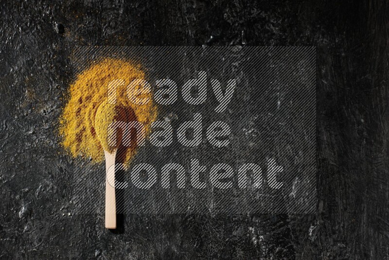 A wooden spoon full of turmeric powder on textured black background