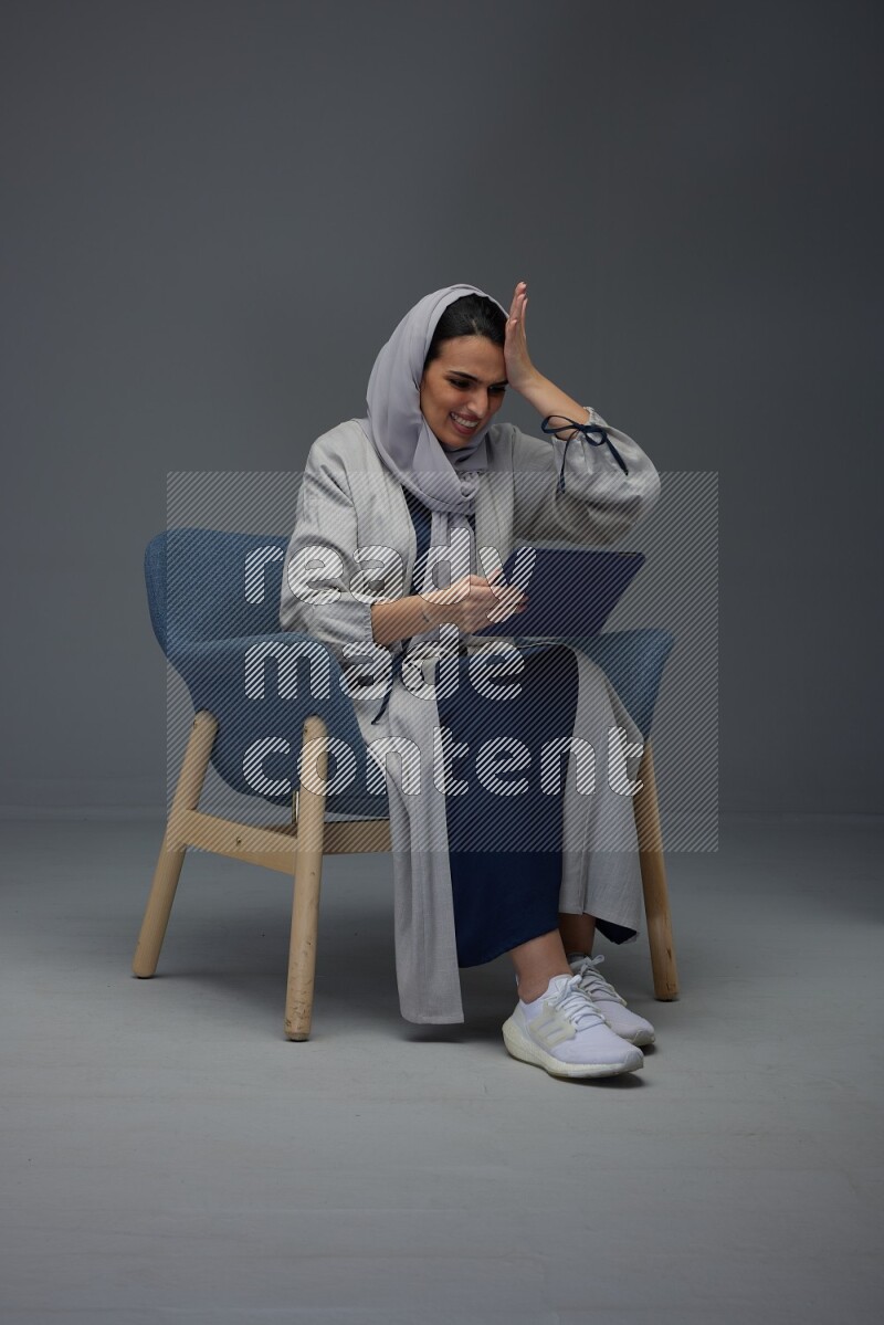 A Saudi female wearing a light gray Abaya and head scarf sitting on a dark grey chair while holding her iPad on a grey background