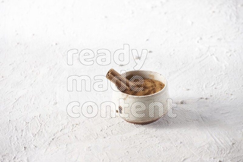 Ceramic beige bowl full of cinnamon powder with a cinnamon stick on a textured white background