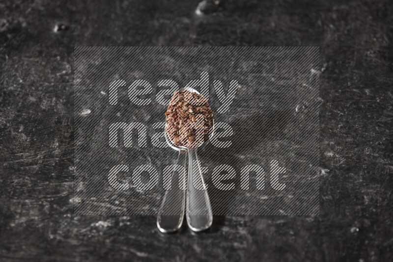 2 metal spoons full of flaxseeds on a textured black flooring