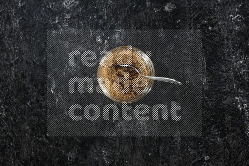 وعاء زجاجي للتوابل وملعقة معدنية ممتلئين ببودرة الكمون على خلفية سوداء