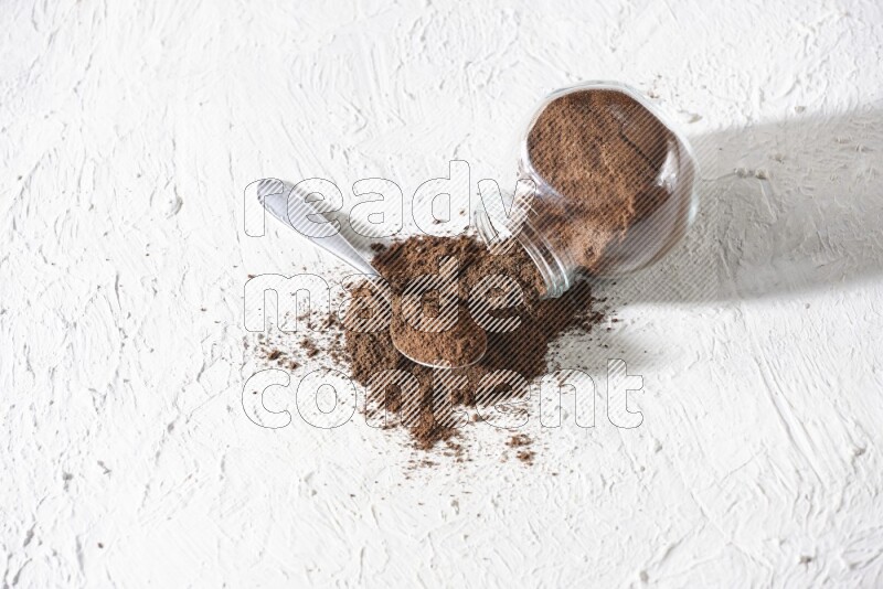 وعاء زجاجي مقلوب وملعقة معدنية ممتلئان ببودرة القرنفل وتناثرت البودرة من الوعاء مع حبوب القرنفل المنتشرة على أرضية بيضاء