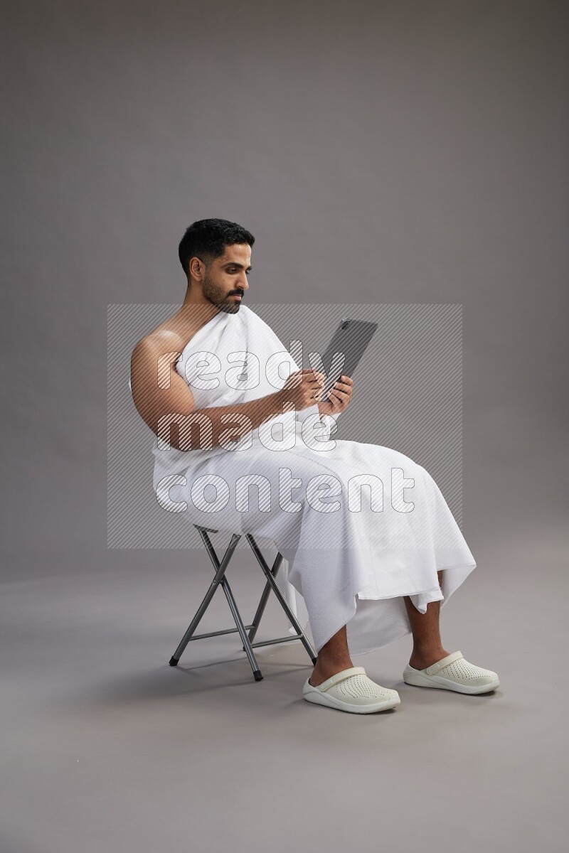 A man wearing Ehram sitting on chair holding a tablet on gray background