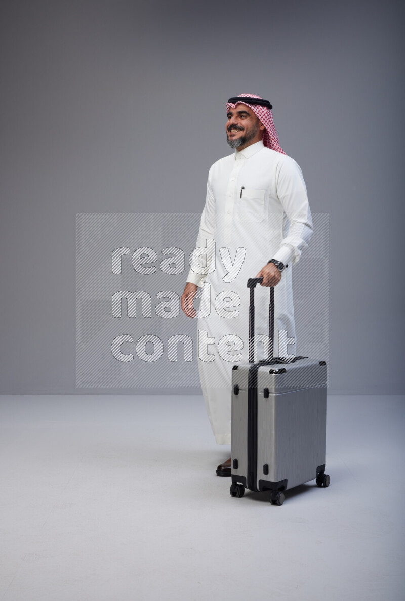 Saudi man wearing Thob and red Shomag standing holding Travel bag on Gray background