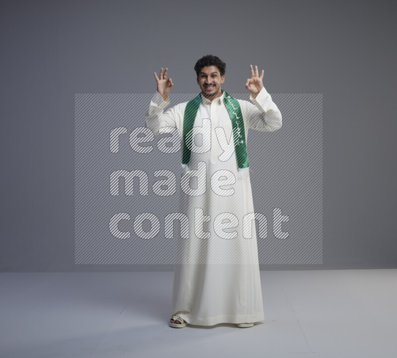 A Saudi man standing wearing thob and Saudi flag scarf on gray background