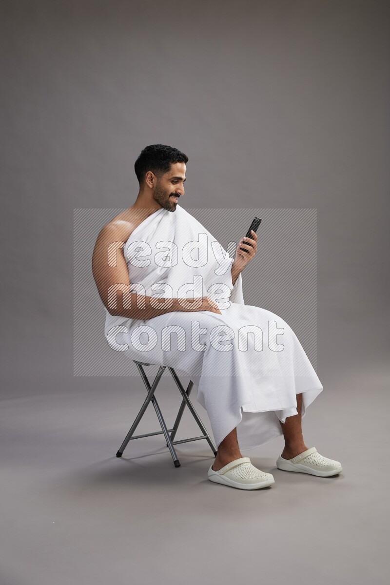 A man wearing Ehram sitting on chair texting on phone on gray background