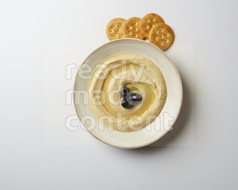 Hummus in a pottry plate garnished with black olives on a white background
