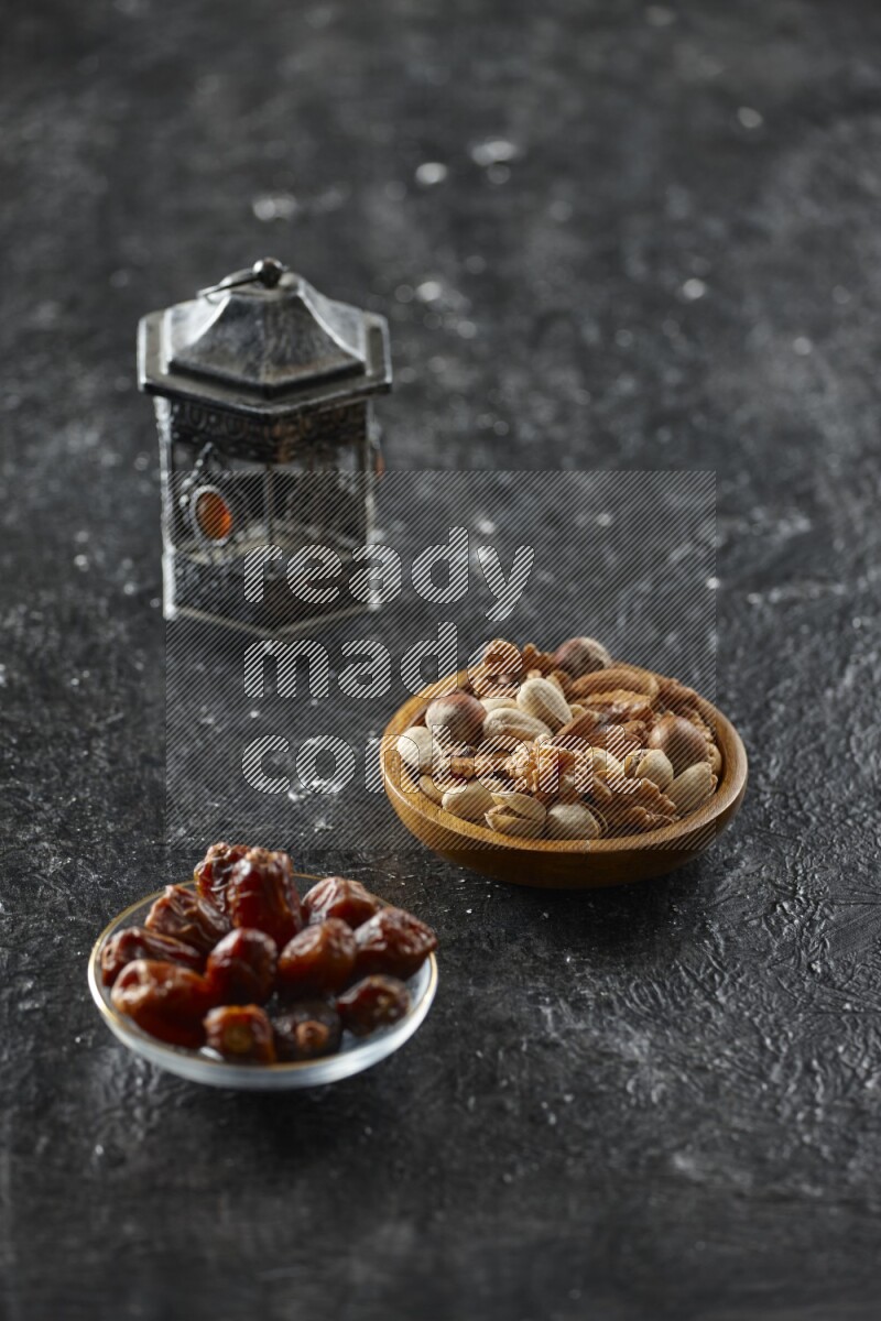 A silver lantern with different drinks, dates, nuts, prayer beads and quran on textured black background