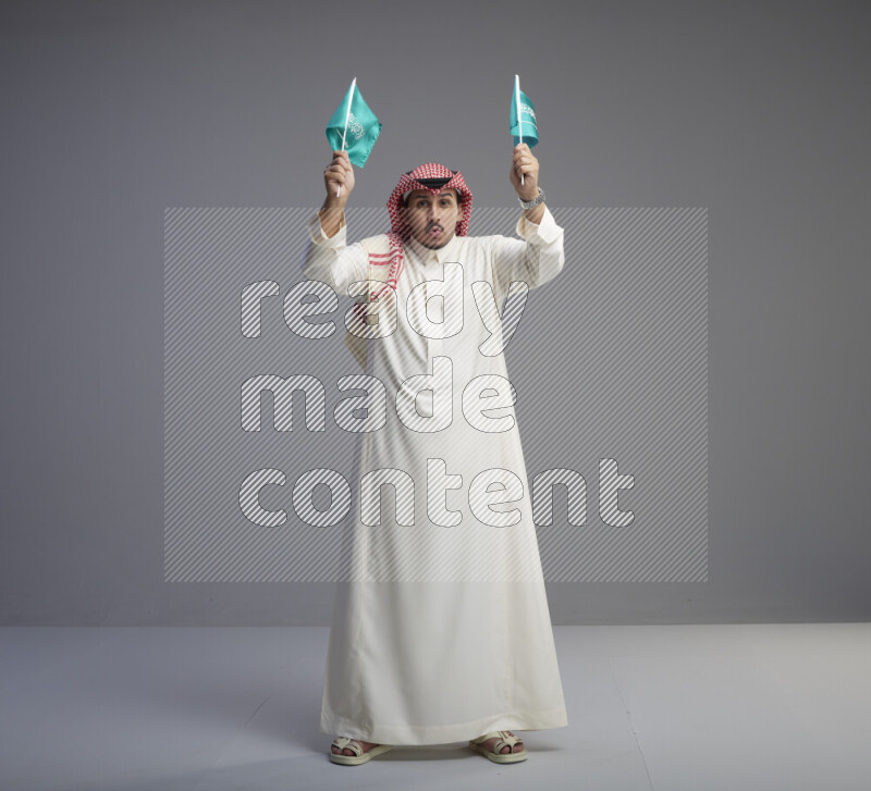 A Saudi man standing wearing thob and red shomag raising small Saudi flag on gray background