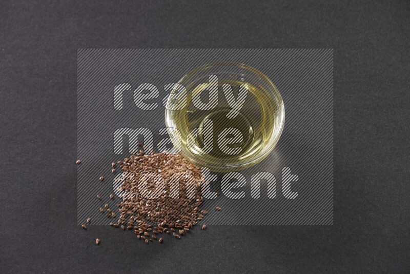 A glass bowl full of flaxseeds oil with some of flaxseeds beside it on a black flooring