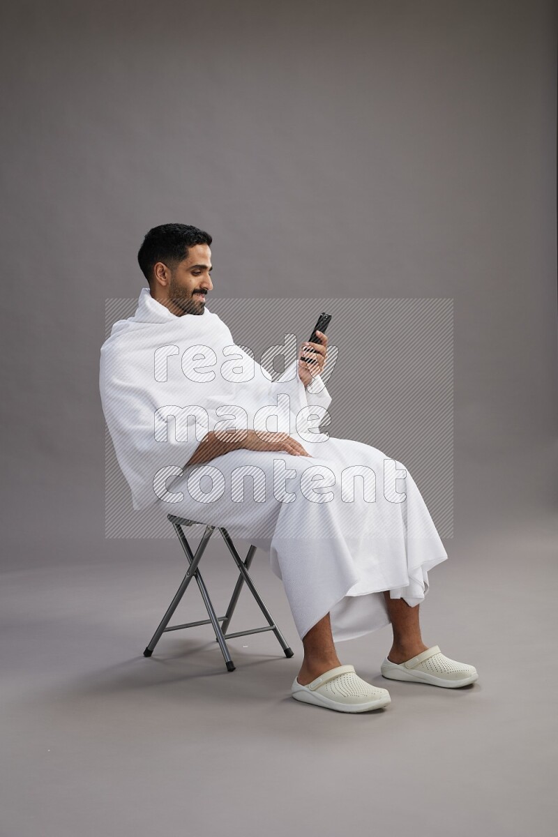 A man wearing Ehram sitting on chair texting on phone on gray background