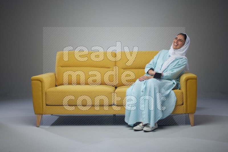 A Saudi woman wearing a light blue Abaya and a white head scarf sitting on a yellow sofa and talking in the phone while making different poses eye level on a grey background