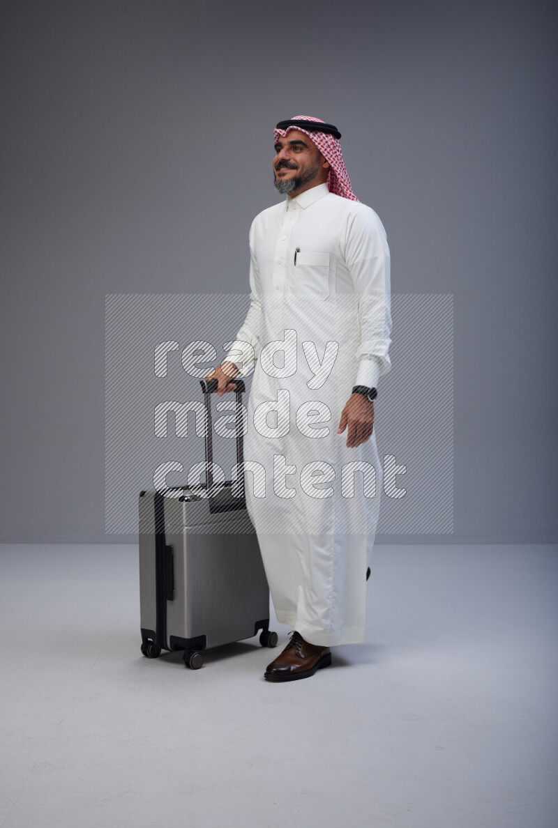 Saudi man wearing Thob and red Shomag standing holding Travel bag on Gray background