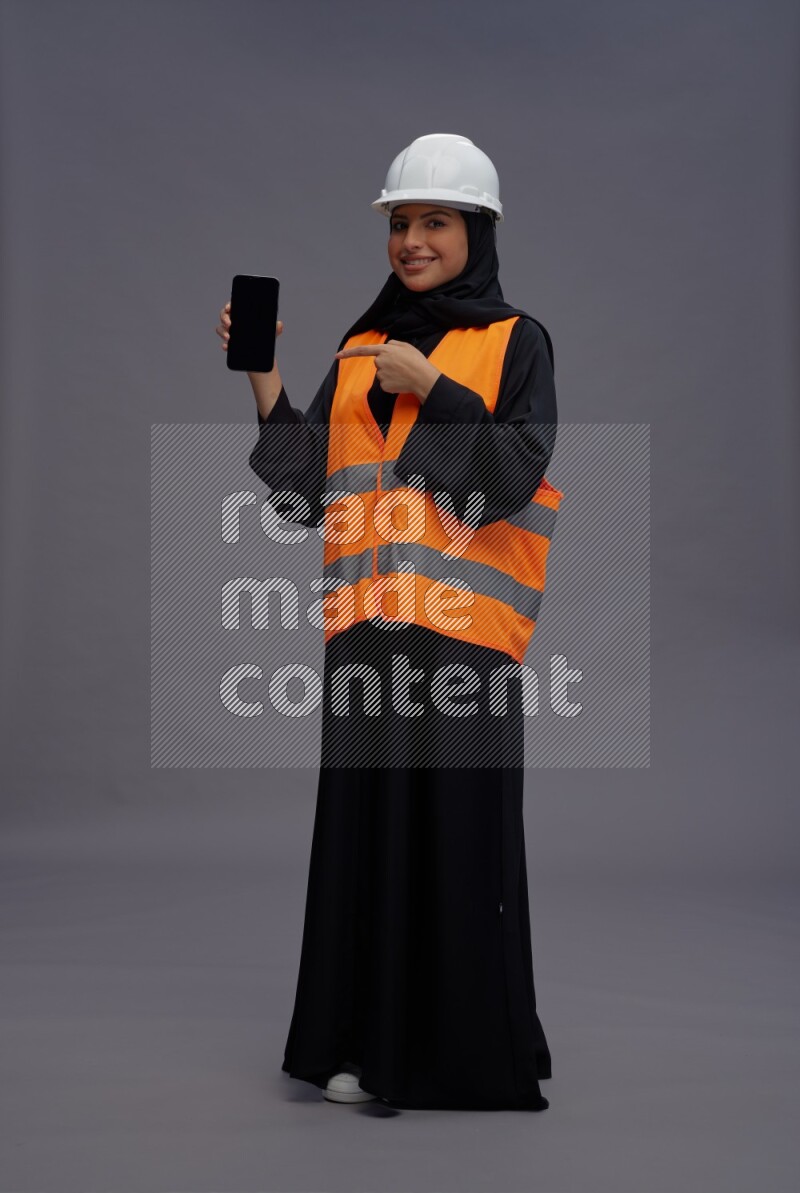 Saudi woman wearing Abaya with engineer vest standing showing phone to camera on gray background