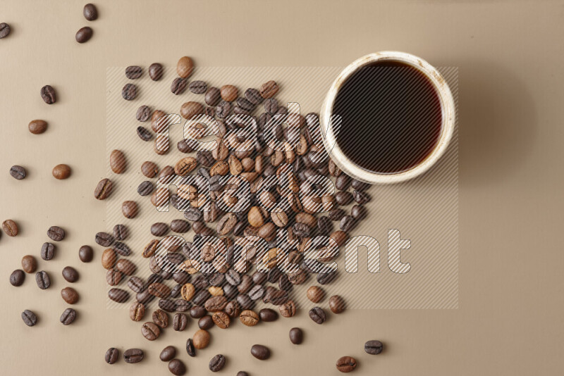 A beige pottery cup of coffee surrounded by roasted coffee beans on beige background