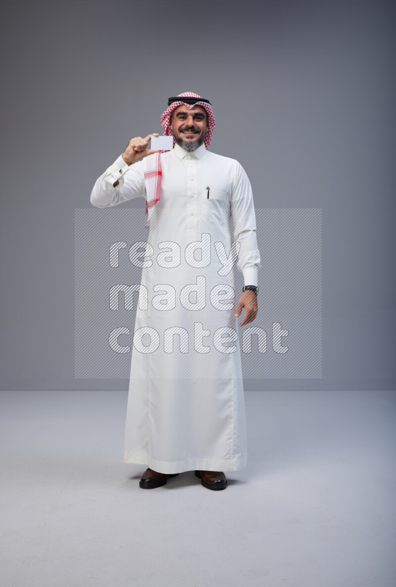 Saudi man Wearing Thob and red Shomag standing holding ATM card on Gray background