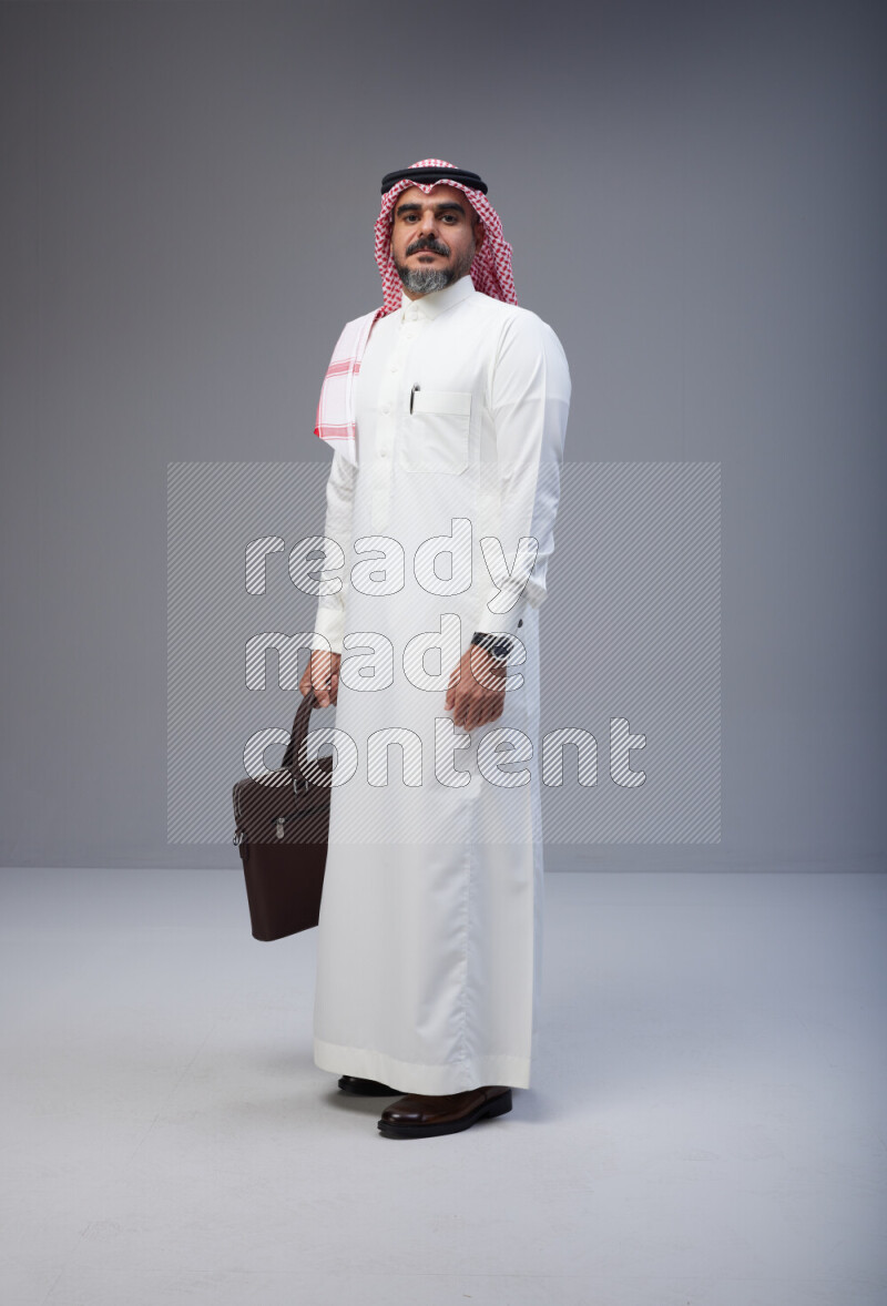 Saudi man Wearing Thob and red Shomag standing holding bag on Gray background