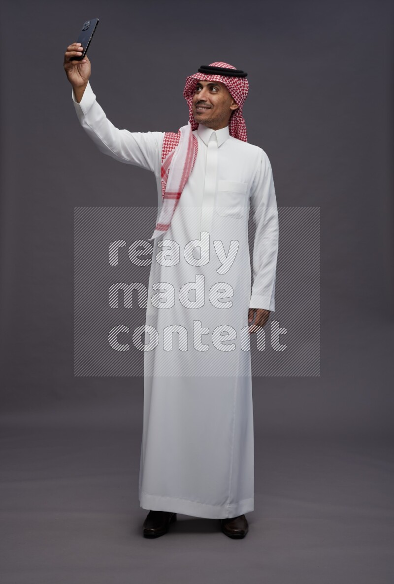 Saudi man wearing thob and shomag standing taking selfie on gray background