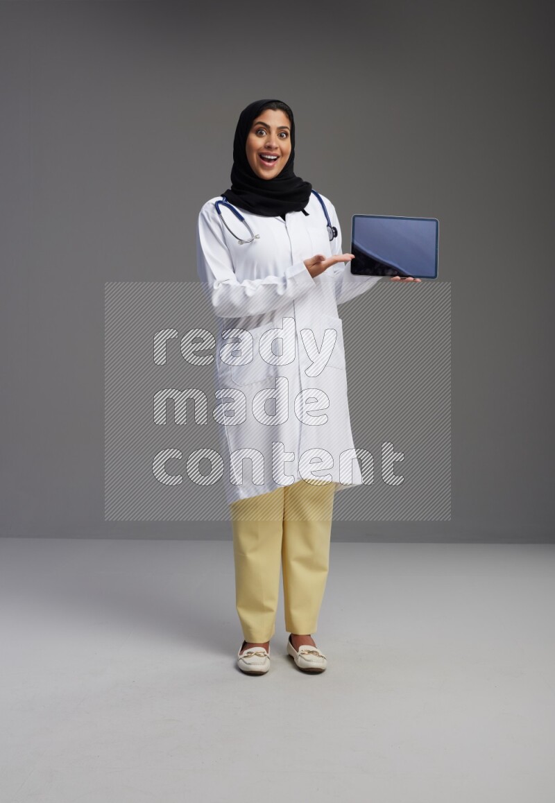 Saudi woman wearing lab coat with stethoscope standing showing tablet to camera with sign in the back on Gray background