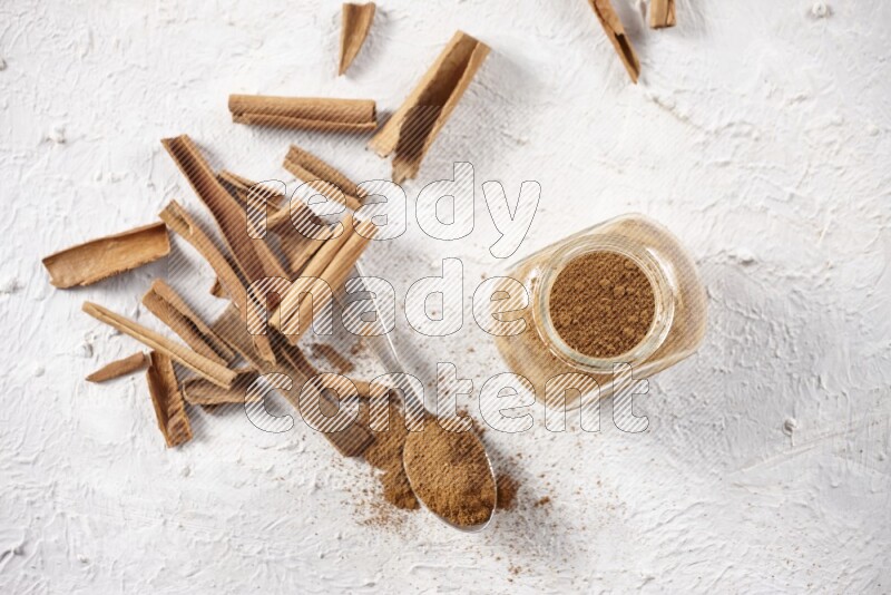 Herbal glass jar full cinnamon powder and a metal spoon surrounded by cinnamon sticks on a white background